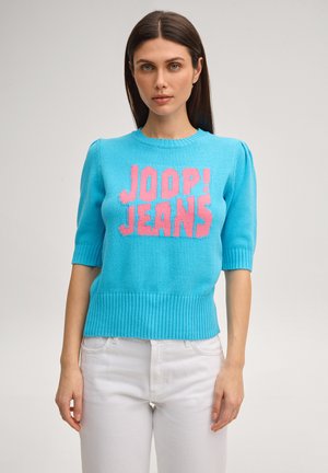 Kobieta z prostymi brązowymi włosami nosi jasnoniebieski krótki dzianinowy top z napisem „JOOP! JEANS” różowymi literami, w połączeniu z białymi spodniami.