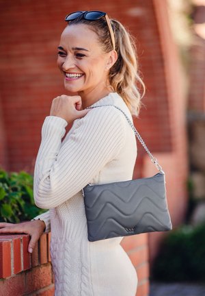 Graue, gesteppte Ledertasche mit einem silbernen Kettenriemen. Sie hat ein wellenförmiges Design und einen Reißverschluss. Lässig zu einem hellen Pullover getragen.