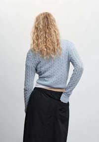 Personne aux longs cheveux blonds bouclés portant un pull en maille torsadée bleu clair et une jupe noire, debout, une main dans la poche, de dos.