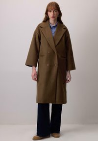 Vrouw staand in een bruin oversized dubbel-gebreid jas, een blauw gestreept shirt, donker wijde broek en lichtbruine schoenen voor een effen achtergrond.