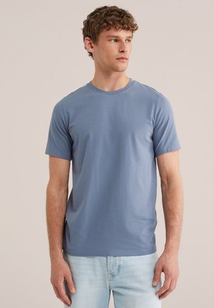 Noor mees lokkis juustega, kandes lihtsat sinist lühikese varrukaga T-särki ja helepesu teksapükse, vaadates veidi kõrvale, neutraalne taust.