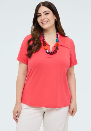 Lachende vrouw met lang donker haar draagt een koraalroze shirt met korte mouwen, een kleurrijke kralenketting en witte broek tegen een effen lichte achtergrond.