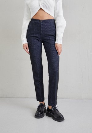 Mujer con pantalones ajustados azul marino con patrón de chevron, suéter de punto blanco corto y zapatos negros gruesos con cordones contra una pared gris.