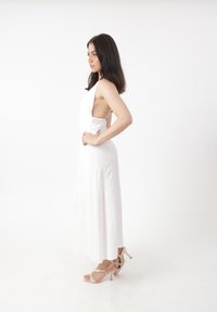 Abito lungo bianco con scollo all' Americana, schiena scoperta e silhouette fluida. Indossato con scarpe con tacco beige. Tessuto morbido.