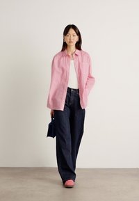 Pink striped button-up shirt, white tank top, dark wide-leg jeans, and pink shoes; holding a small blue handbag. Simple backdrop.