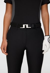 Czarny pleciony pasek z prostokątną srebrną klamrą. Model nosi czarną bluzkę i białą rękawiczkę golfową, prezentując gładki materiał i dopasowany krój.