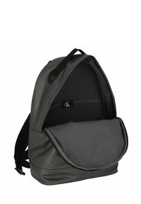 Zaino in tessuto grigio scuro, dotato di un compartimento principale con patta, chiusura a zip e spallacci regolabili. Tasca interna visibile.