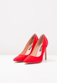 Chaussures à talons hauts en daim rouge avec des pointes effilées, ornées de clous en argent à l'arrière. Dotées d'une doublure intérieure lisse.