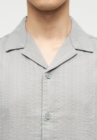 Κοντινό πλάνο ατόμου που φοράει ανοιχτό γκρι υφασμάτινο πουκάμισο με γιακά με εγκοπή και δύο εμφανείς κουμπωμένες κουμπιές.