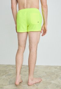 Pantaloni scurți de înot neon verzi, cu un tiv elastic, croi scurt și un singur buzunar pe spate. Textură netedă, design simplu, purtați desculți pe o podea din piatră.