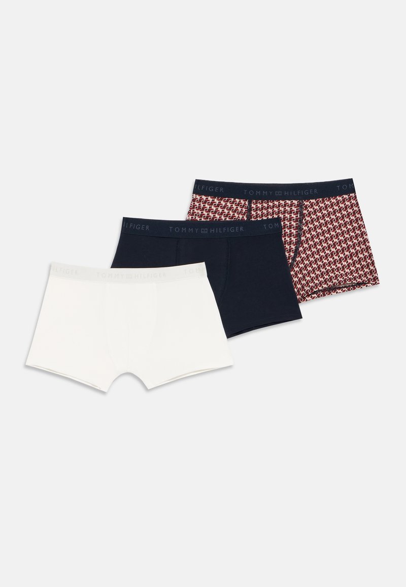 Tri moške spodnje boksarice v beli, enobarvni mornarsko modri in rdeči z vzorcem, vsake s pasom z logotipom Tommy Hilfiger, položene na ravni beli podlagi.