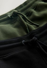 Zwei Paare von Jogginghosen: eine olivgrün mit einem strukturierten Bund und Kordelzug; eine einfarbig schwarz mit einem glatten Bund und Kordelzug.