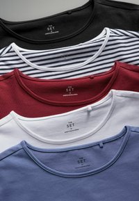 Quattro t-shirt impilati: nero, bianco, bordeaux e blu. Ognuno ha un collo a giro, una morbida trama di cotone e l'etichetta "THE SET" sul retro.