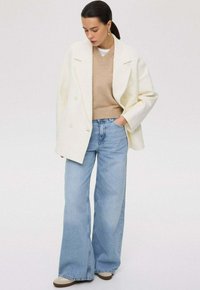 Witte dubbelzijdige jas over een beige v-hals trui, gecombineerd met lichtblauwe brede jeans en crèmekleurige sneakers, op een neutrale achtergrond.