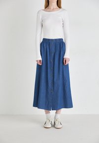 Gonna maxi in denim con bottoni frontali, cintura elastica e tasche laterali. Indossata con un top bianco a maniche lunghe a coste e sneakers.