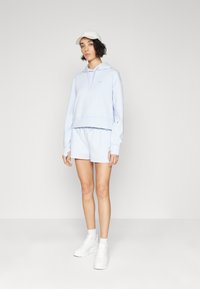 Femeie îmbrăcată cu o bluză ușor albastră, pantaloni scurți asortați, adidași albi, șosete și o șapcă albă, stând în fața unui fundal simplu deschis.