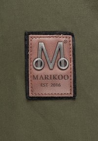 Olivinės spalvos audinyje yra odinė etikete su įspaustu sidabriniu "M" logotipu, supa juodas siūlas. Tekstas skaito "MARIKOO EST 2016."