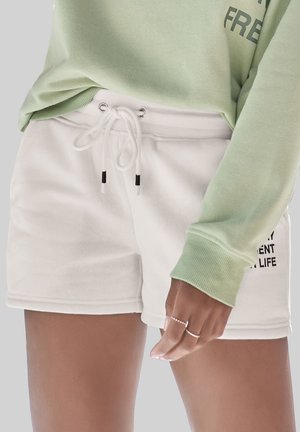 White cotton shorts with an elastic waistband, drawstring with black tips, and side pockets. Paired with a light green, long-sleeve top.