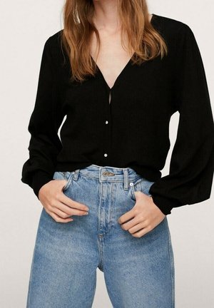 Vrouw draagt een zwarte V-hals blouse met knopen en lange mouwen, ingestopt in een lichtblauwe high-waisted spijkerbroek, handen in de voorzakken.