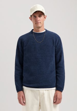 Navy blue textured sweater with a crew neckline and long sleeves, layered over a white shirt. Worn with a light-colored cap and chain necklace.