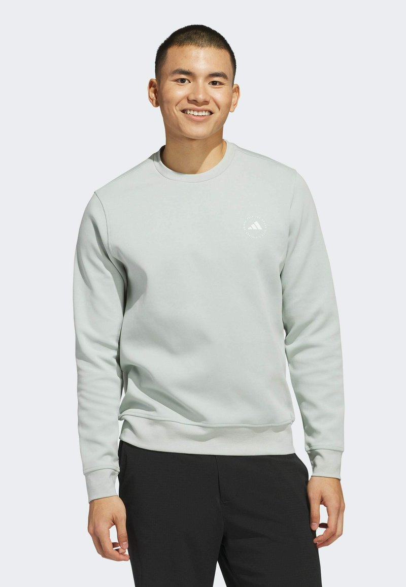Felpa grigia chiara con scollatura rotonda, polsini e orlo a coste. Presenta un logo bianco sul petto e una texture liscia. Indossata con pantaloni neri.