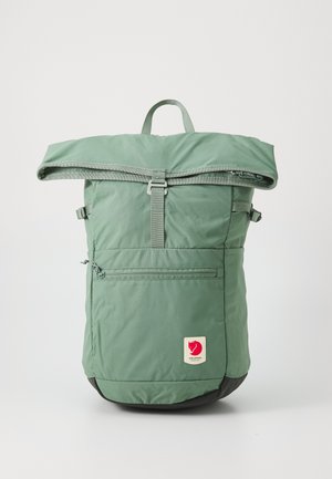Grüner Stoff-Rucksack mit einem Roll-Top-Design, einer vorderen Reißverschlusstasche, verstellbaren Trägern und einem schwarzen Boden. Mit einem roten Logo-Patch.
