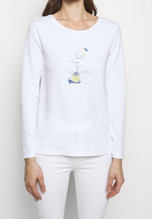 Femme portant une chemise blanche à manches longues avec une illustration marine d'un panneau indicateur, d'une mouette et d'une valise jaune sur le devant.