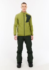 Man in een groene zip-up jas en donkergroene broek staat op een witte achtergrond, recht vooruit kijkend met ontspannen armen.