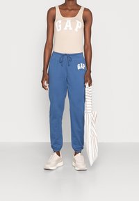Débardeur beige avec logo "GAP", pantalon de survêtement bleu avec logo, et baskets claires. Tenant un sac rayé ; tenue décontractée et confortable.