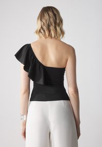 Černý top na jedno rameno s volánkovým akcentem, vyrobený z hladké látky, spárovaný s bílými kalhotami. Minimalistický design zvýrazňuje siluetu.