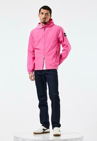 Letvægts pink jakke med hætte, lynlås lukning og elastiske manchetter. Båret med mørke jeans og hvide sneakers på en marmorpodest.
