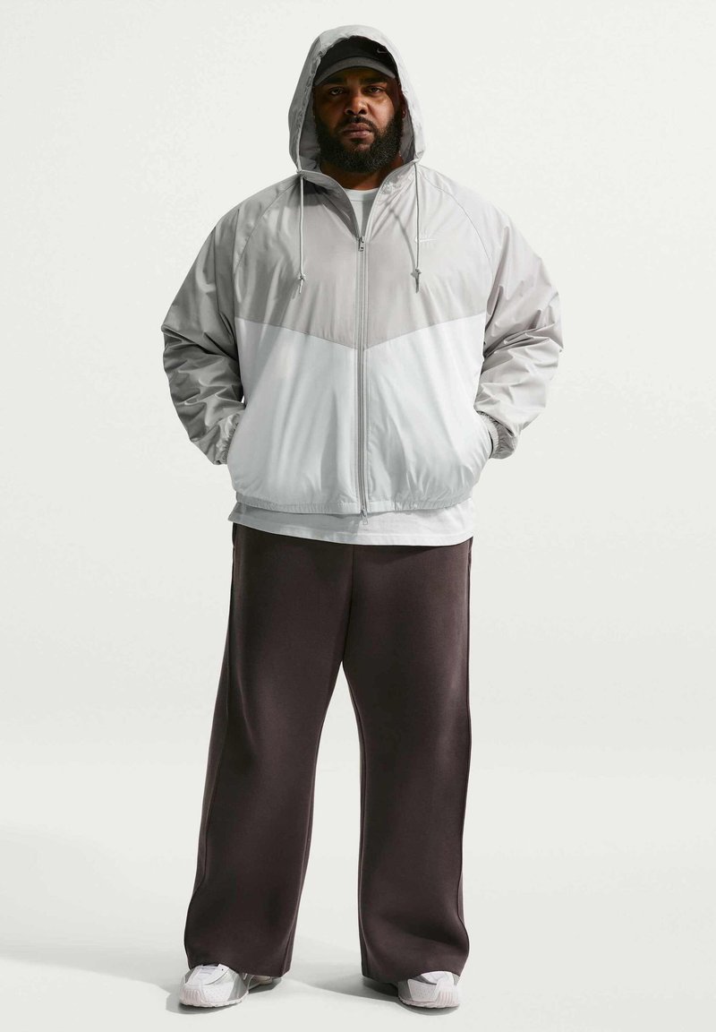 Hombre con barba que lleva un cortavientos gris con capucha, gorra negra, pantalones anchos oscuros y zapatillas blancas, de pie con las manos en los bolsillos.