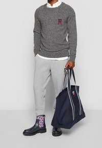 Grauer Strickpullover mit Logo, weißes Hemd mit Kragen, graue Jogginghose, schwarze Stiefeletten mit gemusterten Akzenten und eine marineblaue Tragetasche.