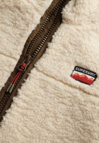 Giacca in pile color crema con zip marrone e cordoncino rosso, dotata di una patch del logo Superdry con design montano sul petto.