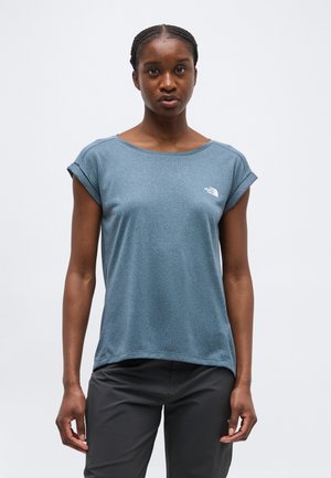 Mujer con el cabello trenzado, vestida con una camiseta azul de manga corta y pantalones oscuros, de pie contra un fondo claro y liso.