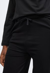 Črne hlače za prosti čas z elastičnim pasom in vrvico za zategovanje, z gladko, mehko teksturo. Delno vidne pod črnim zgornjim delom.