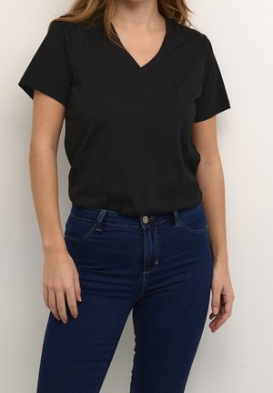 Kvinde iført en ensfarvet sort T-shirt med korte ærmer og V-hals samt mørkeblå tætsiddende jeans, stående op ad en hvid væg.