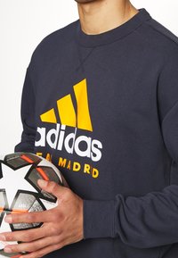 Sudadera azul marino con el logo blanco de "adidas" y acentos amarillos. El diseño incluye un cuello redondo y puños acanalados. La persona sostiene un balón de fútbol en blanco y negro.