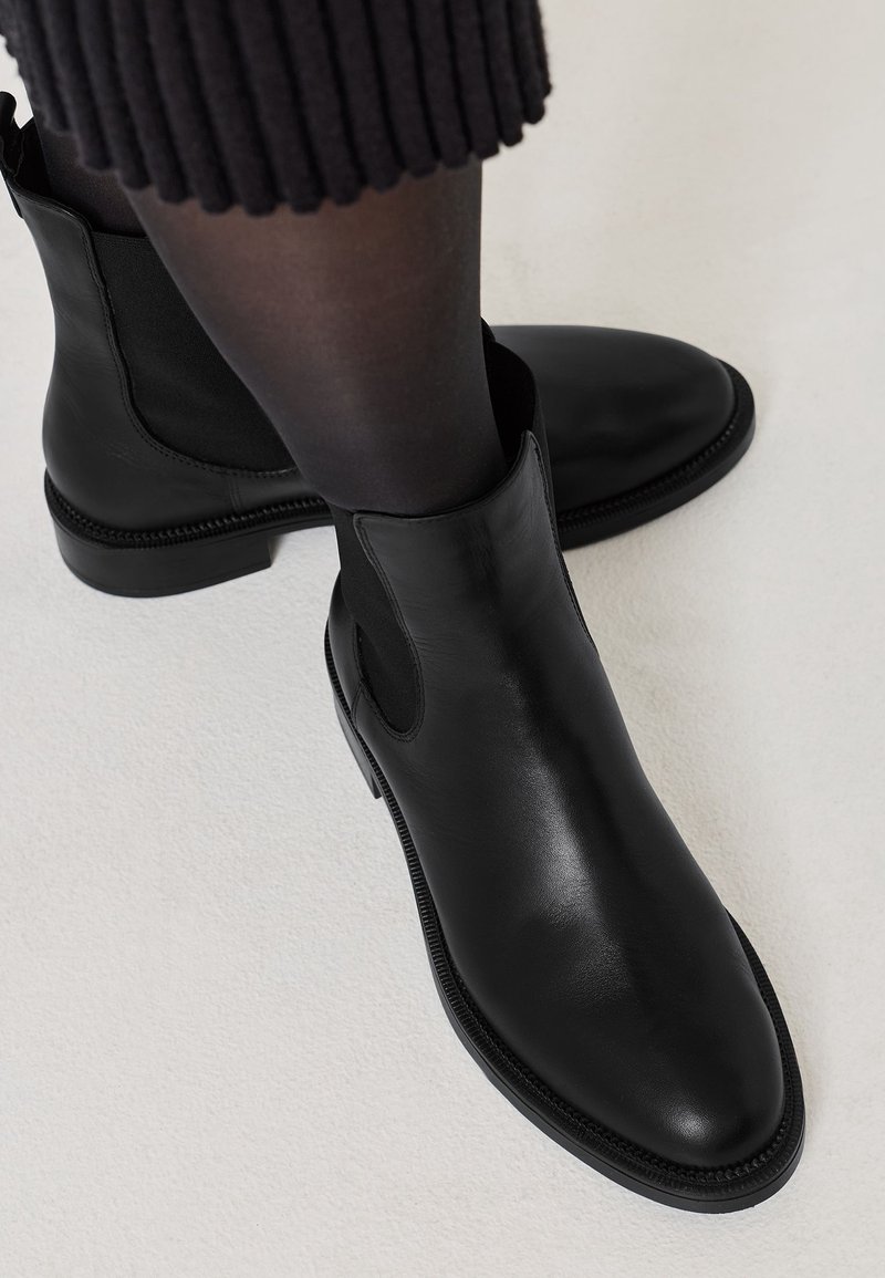 Bottines en cuir noir portées avec des collants noirs transparents et une jupe côtelée foncée sur un fond de sol clair.