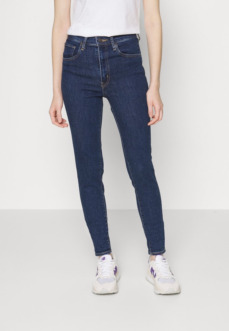 Jean taille haute bleu foncé avec une coupe slim, cinq poches et des détails de couture visibles. Assortis à des baskets blanches et violettes.