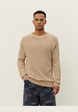 Beige ribbed knit sweater with long sleeves, round neckline, and textured pattern; worn with patterned blue pants featuring various designs.