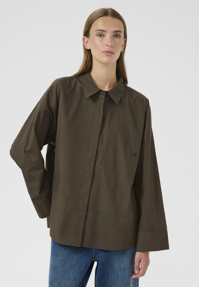 Loose-fitting brown shirt with a collar, long sleeves, and a button detail on the left chest, paired with blue jeans.