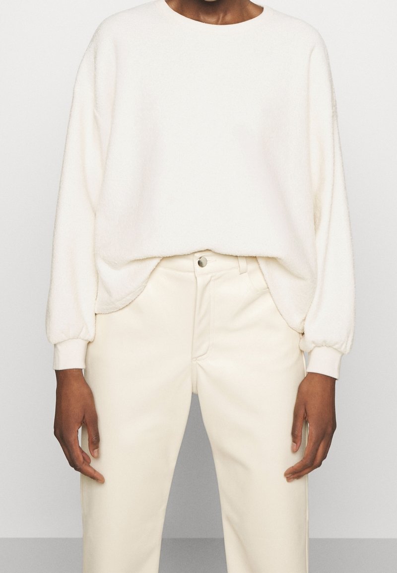 Pullover bianco in pile con collo tondo e maniche lunghe a sbuffo, abbinato a pantaloni bianchi su misura con chiusura frontale a bottoni.