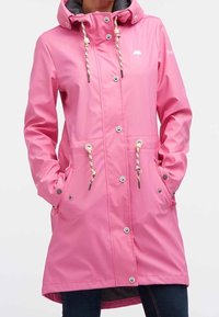 Nainen, jolla on pitkä vaaleanpunainen sadeviitta hupulla, napilla ja vetoketjulla, ja kädet etutaskuissa, yhdistettynä tummiin farkkuihin.