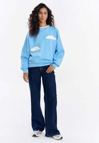 Niebieska bluza z białymi wzorami chmur i napisem. Miękki materiał polarowy, luźny krój, zestawiona z ciemnymi dzwonowatymi spodniami dżinsowymi i sneakersami.