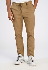 Helle braune tapered Pants mit einem Kordelzugbund und zwei Vorder Taschen. Getragen mit weißen Sportschuhen, die farbenfrohe Akzente aufweisen.