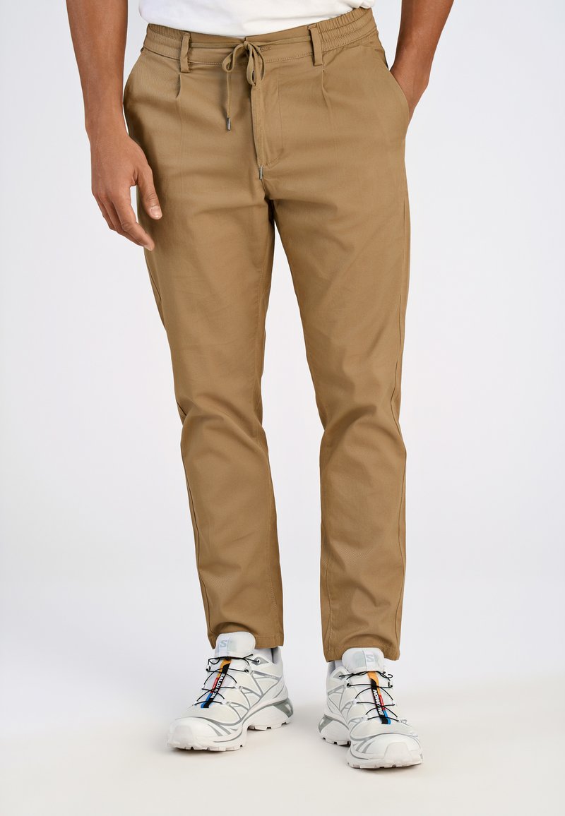 Helle braune tapered Pants mit einem Kordelzugbund und zwei Vorder Taschen. Getragen mit weißen Sportschuhen, die farbenfrohe Akzente aufweisen.