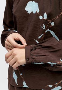 Blouse marron avec une finition texturée arborant des imprimés floraux bleu clair. Manches longues avec poignets froncés et un détail de bouton au niveau du poignet.