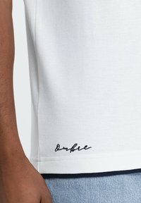 Polo bianco in tessuto testurizzato, con un logo ricamato nero all'orlo e un accento blu navy a contrasto nella parte inferiore.