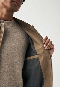 Veste en daim beige avec une doublure intérieure grise. Le col est doux et le matériau a une texture lisse. Le modèle porte un pull beige en dessous.